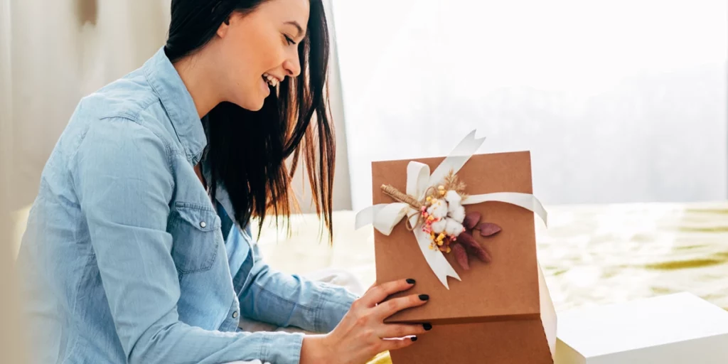 Side view of happy young woman sit on the bed unpacking a beautifully packaged parcel from an online shop.