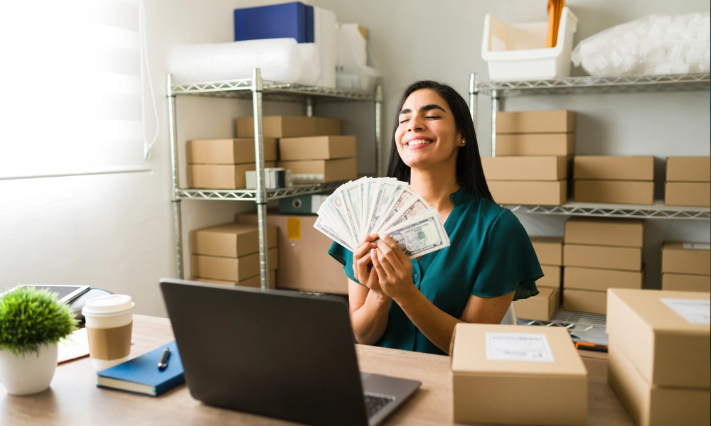 business women sitting at a desk, holding a fan of money with a happy look on her face.